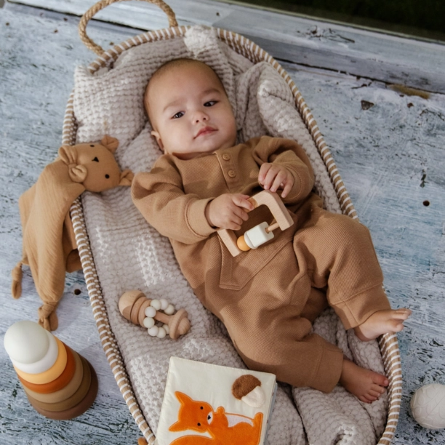 Baby lying in a cozy basket surrounded by neutral baby toys including a wooden rattle, lovey, and silicone stacking tower.