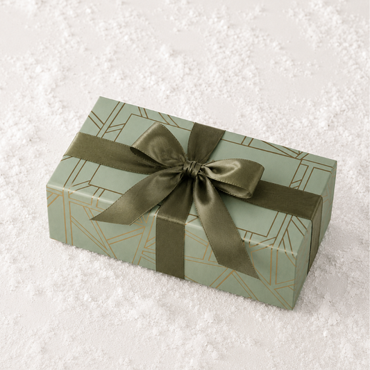 A rectangular gift box wrapped in sage green paper with a geometric gold line pattern and tied with an olive green satin ribbon, placed on a white textured surface.