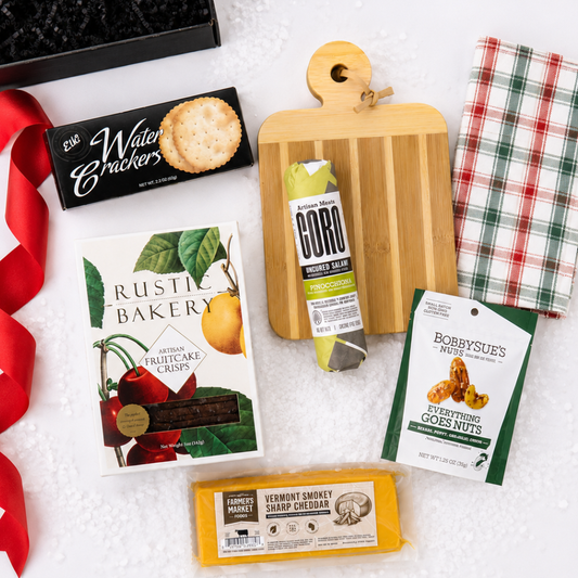 A curated gift basket or snack assortment laid out on a white textured surface, including a black box of Water Crackerz, a box of Rustic Bakery artisan fruit crisps, a package of Gordo smoked turkey, a bag of Bobbysues' New York Everything Goes Nuts, a block of Vermont Smokey Sharp cheddar cheese, a wooden cutting board, a red-and-white plaid cloth, and a red ribbon.