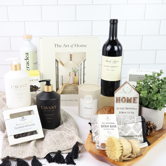 Collection of home products including a bottle of wine, candles, and soaps on a white surface with a 'The Art of Home' book. Stunning luxury kitchen gift basket fro Seattle delivery.