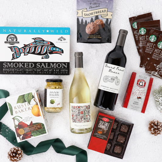 A curated gift basket flat lay on a white textured background, featuring a smoked salmon box, a bottle of white wine, a bottle of red wine, hot chocolate packs, a shortbread cookie bag, a box of Rustic Baker cookies, a jar of salami, chocolates, and a green ribbon.