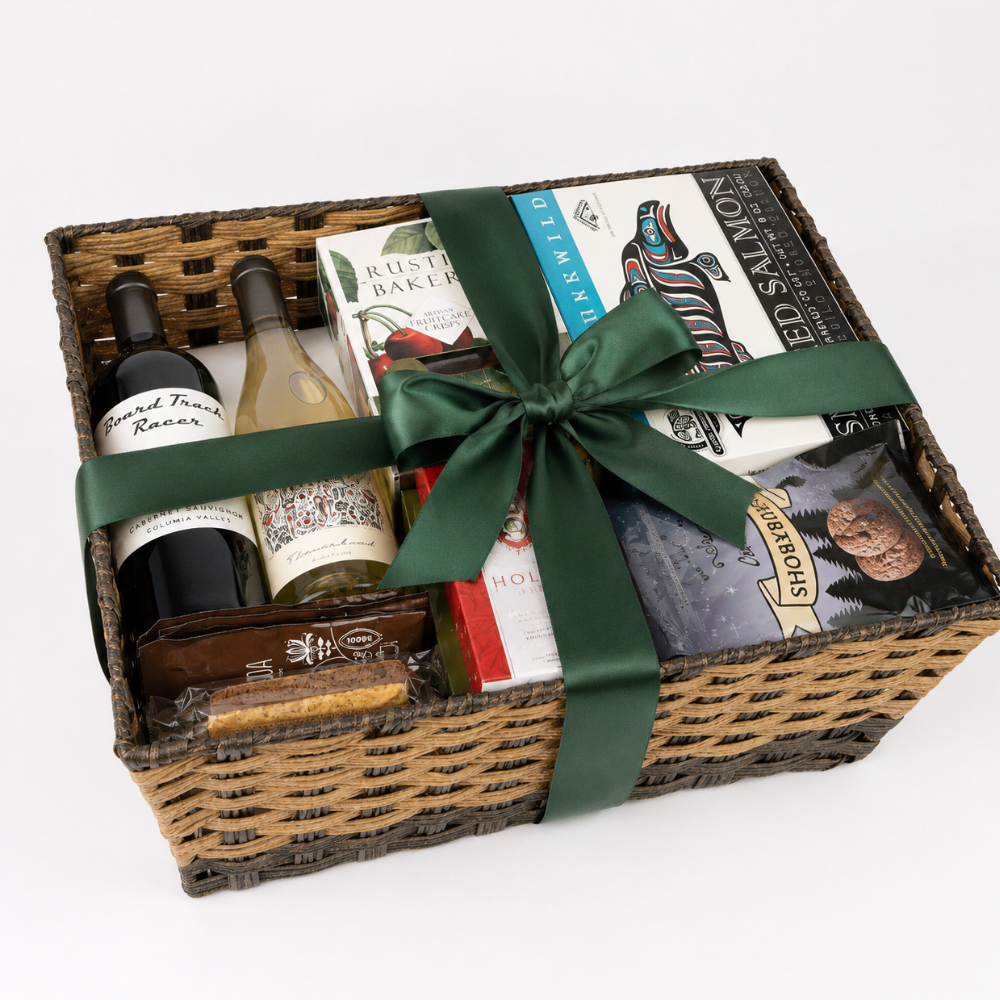 A wicker gift basket filled with assorted gourmet items, tied with a dark green ribbon. Visible contents include a bottle of red wine, a bottle of white wine, a box of rustic bakeries, a book, and packaged snacks.