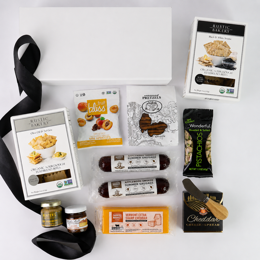 A curated gourmet gift basket flat-lay featuring various food products from brands like Rustic Bakery, Bliss, and Peddler's on a clean white background with a black ribbon.