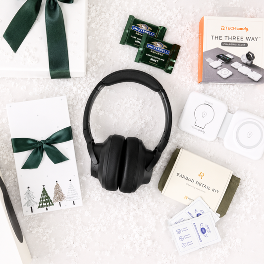 Tech Candy 'The Three Way' earbud gift set arranged on a white textured background, featuring black over-ear headphones, earbud cases, earbud detail kit, gift boxes with dark green ribbons, and green packaged candy.