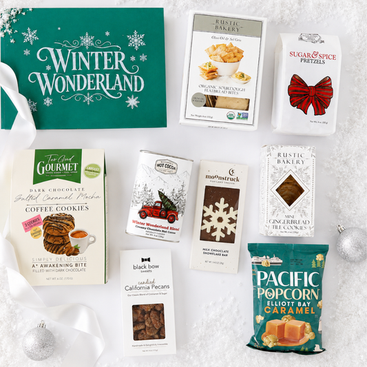 A holiday gift basket assortment of gourmet snacks and cookies, featuring a green 'Winter Wonderland' gift box, Rustic Bakes chocolate chip cookies, sugar and spice pretzels, Gourmet coffee cookies, a can of Oreo chocolate milk cookies, mochapstick marshmallow treats, black bow California pecans, and a bag of Pacific Popcorn Elliott Bay Caramel, arranged on a white textured surface with silver ornaments.