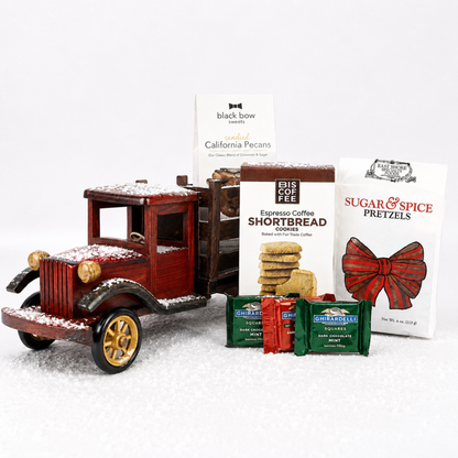 A holiday-themed gift set featuring a red vintage wooden truck with gold wheels and white faux snow, alongside packages of Black Bow Chocolates, Bis in Flee Espresso Coffee Shortbread Cookies, Sugar & Spice Pretzels, and Guatemalco Especial coffee, arranged on a white snowy surface.