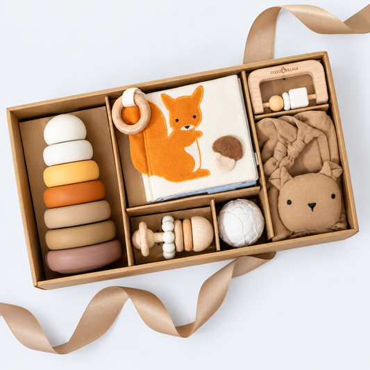 A modern unisex wooden and fabric baby gift set arranged in a kraft cardboard box with ribbon, featuring a colorful ring stack, a fox-themed cloth book, a wooden teething toy, a plush bear, and other soft baby accessories on a white background.