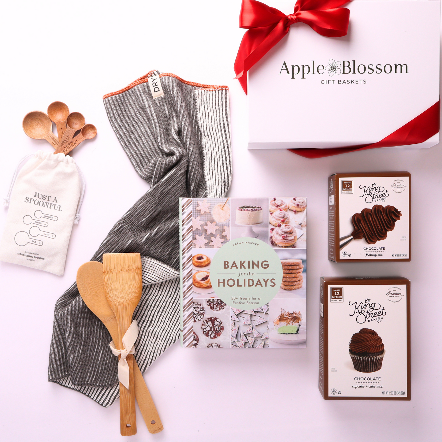 Flat lay of holiday baking gift box with Baking for the Holidays cookbook, chocolate cake mix, frosting mix, kitchen towel, wooden utensils, and measuring spoons, with a 5-star customer review.