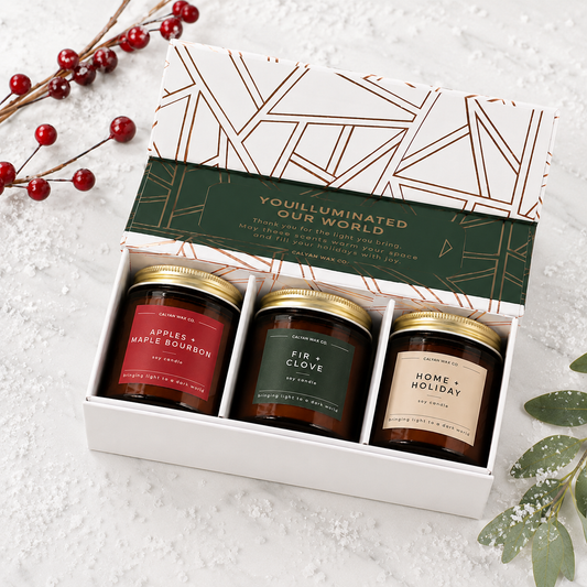 A gift set of three scented candles in a white box with a geometric-patterned lid. The candles have gold lids and labels for 'Apples + Maple Bourbon', 'Fir + Clove', and 'Home + Holiday'.