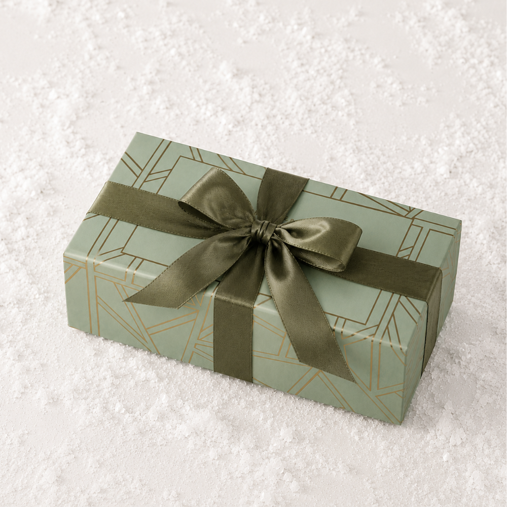 A rectangular gift box wrapped in sage green paper with a geometric gold line pattern and tied with an olive green satin ribbon, placed on a white textured surface.