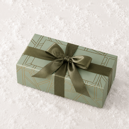 A rectangular gift box wrapped in sage green paper with a geometric gold line pattern and tied with an olive green satin ribbon, placed on a white textured surface.