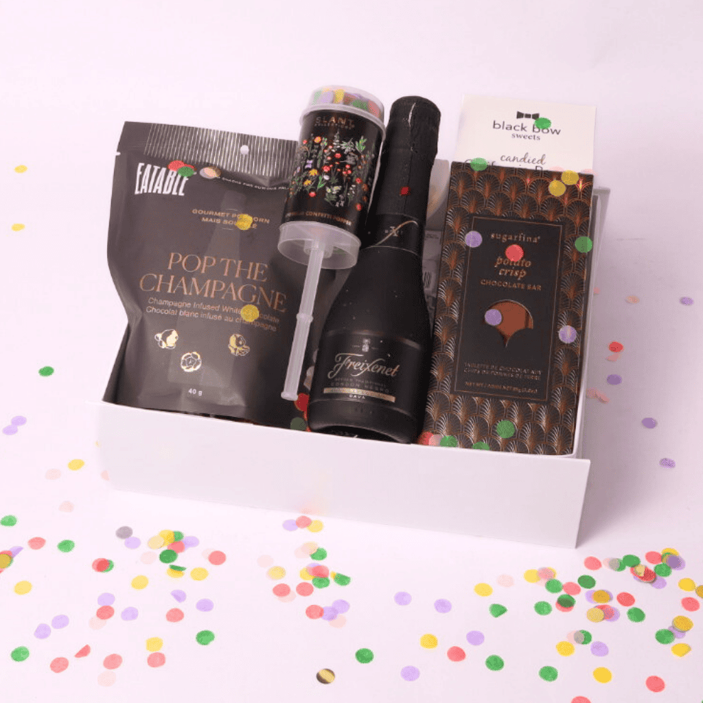 A luxurious celebration gift box featuring champagne-infused popcorn, a mini bottle of Freixenet, a Sugarfina chocolate bar, a confetti popper, and candied nuts, all elegantly arranged in a white box.
