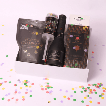 A luxurious celebration gift box featuring champagne-infused popcorn, a mini bottle of Freixenet, a Sugarfina chocolate bar, a confetti popper, and candied nuts, all elegantly arranged in a white box.
