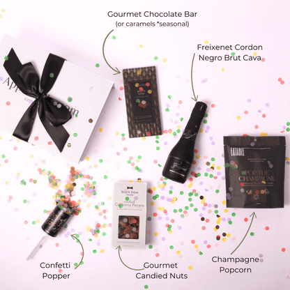 A top-down view of a celebration gift box with all items displayed: champagne popcorn, a Freixenet mini bottle, a gourmet chocolate bar, a festive confetti popper, and candied nuts.