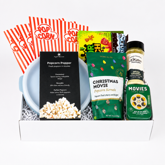 A curated popcorn gift box set in a white tray, featuring red-and-white striped popcorn bags, a blue bowl, a black package labeled Popcorn Popper, a green bag of Christmas Movie popcorn kernels, and spice jars including White Pepper and Movie Night Spices.