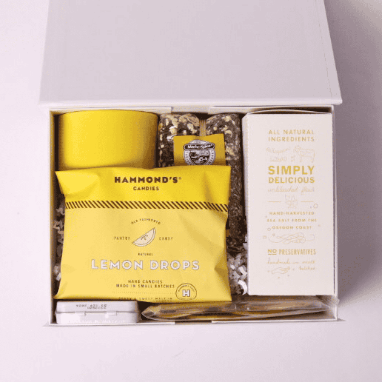 Unboxing image of the A Drop of Joy lemon-themed gift box, beautifully arranged with lemon shortbread cookies, a ceramic yellow teacup, citrus crisps, Seattle Surprise loose tea, lemon drops, and Simply Lemon mints—a perfect care package for her or get well soon gift.