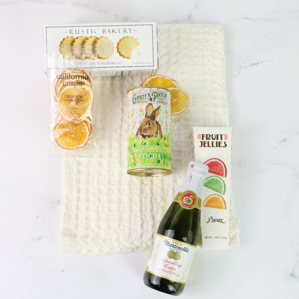 spring gift basket with Assorted items including a bottle of sparkling wine, a can of carrot cake, and a package of orange slices on a textured white surface.
