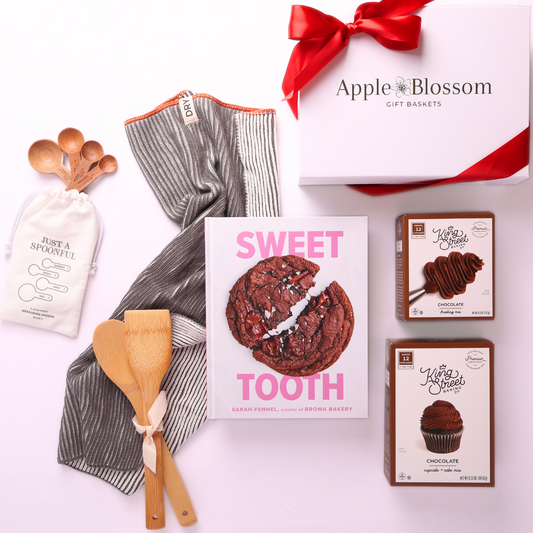 flat lay of gift contents with box and red bow: Let’s Bake housewarming gift box with baking essentials, chocolate mixes, kitchen towel, and measuring spoons in Apple Blossom packaging.