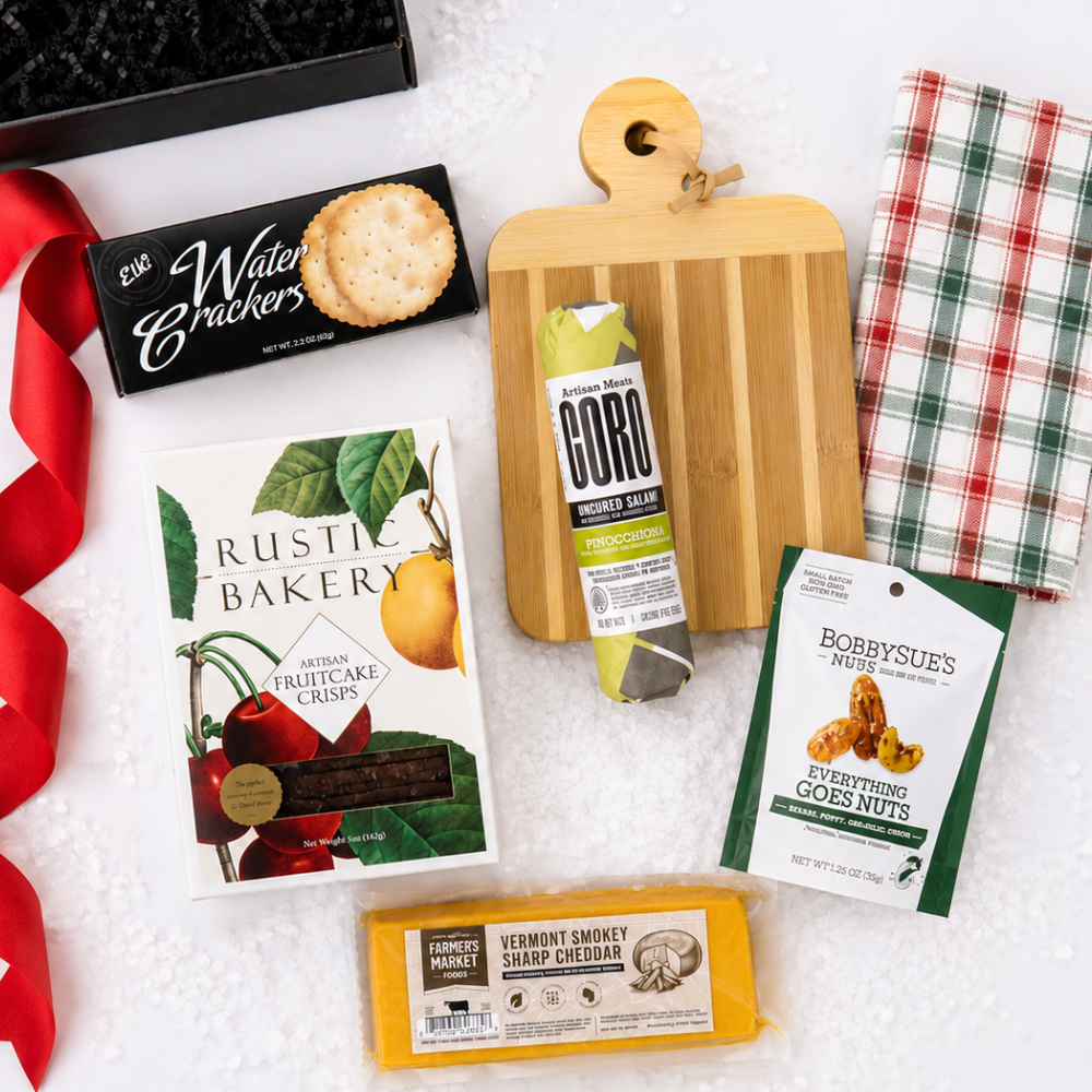A curated gift basket or snack assortment laid out on a white textured surface, including a black box of Water Crackerz, a box of Rustic Bakery artisan fruit crisps, a package of Gordo smoked turkey, a bag of Bobbysues' New York Everything Goes Nuts, a block of Vermont Smokey Sharp cheddar cheese, a wooden cutting board, a red-and-white plaid cloth, and a red ribbon.