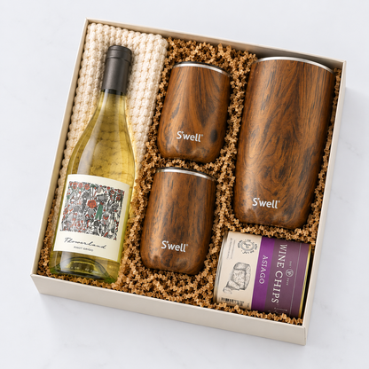 A gift set in a beige box featuring a bottle of white wine, four dark wood Swell insulated tumblers, and a can of Asaago winemakers, arranged in brown crinkle paper on a white background.