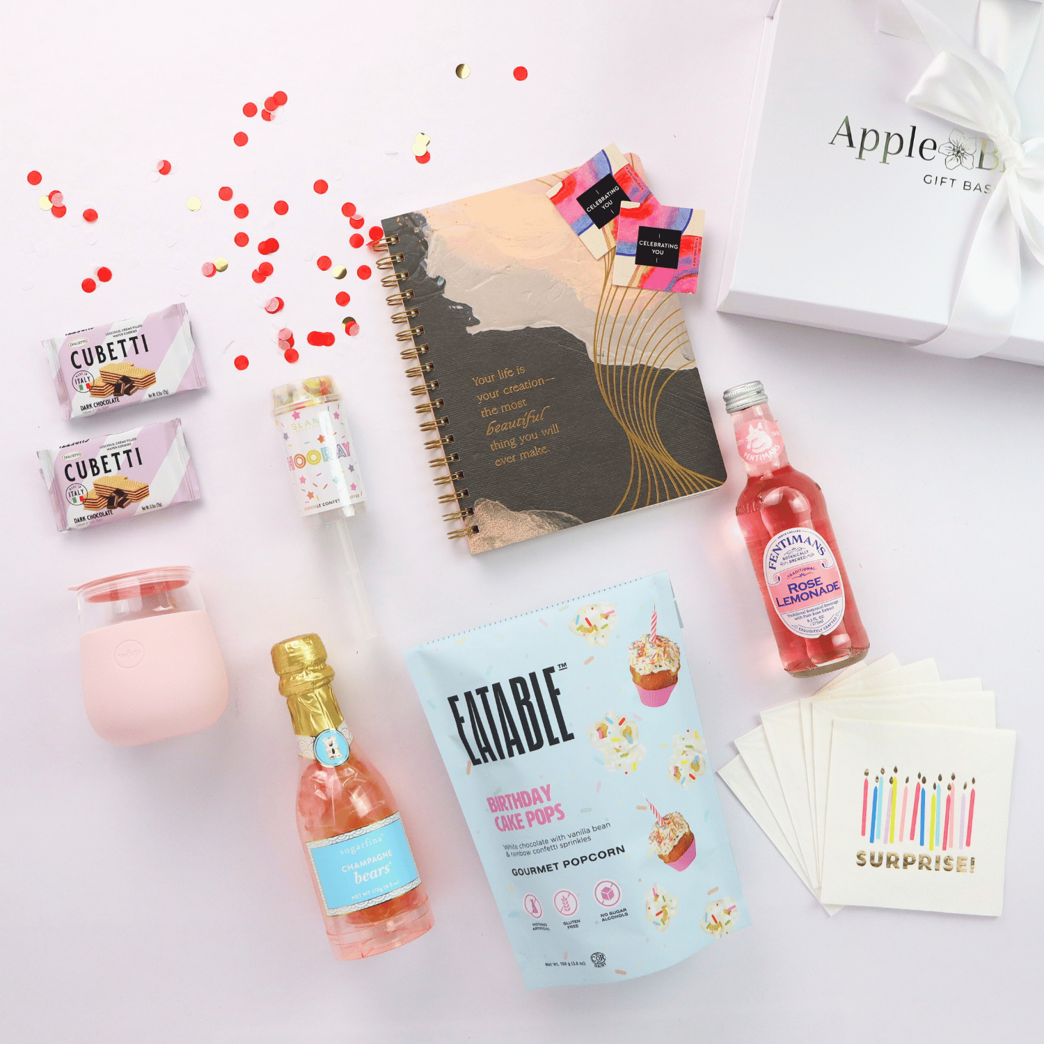 Flat lay of a luxury birthday gift box for her, featuring birthday cake popcorn, rose lemonade, champagne gummy bears, a pink tumbler, and Journal.