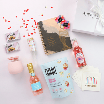 Flat lay of a luxury birthday gift box for her, featuring birthday cake popcorn, rose lemonade, champagne gummy bears, a pink tumbler, and Journal.