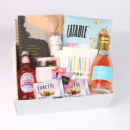 Birthday gift box for her with birthday cake popcorn, lemon shortbread, rose lemonade, champagne gummy bears, a pink "Happy Vibes" tumbler, a party game, and festive accessories.