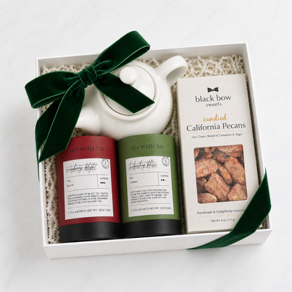 A white gift box filled with tea and sweets, tied with a dark green velvet bow. The box contains a white teapot, two tea tins labeled 'Tea With Tai' in red and green, and a box of 'Black Bow Sweets' featuring 'Candied California Pecans'.