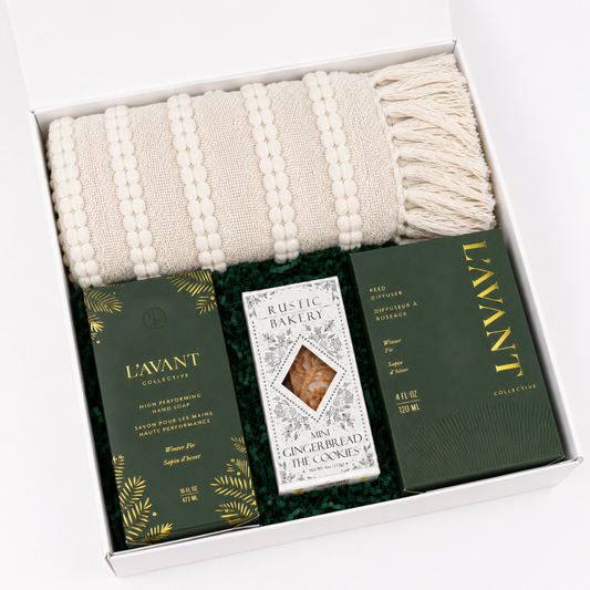 A curated gift set in a white box featuring a cream knit throw blanket with beaded stripes and fringe, a bar of L'Avant Rose Petal soap, a bag of L'Avant Longerbread & The Cookies, and a green L'Avant Reed Balm box.