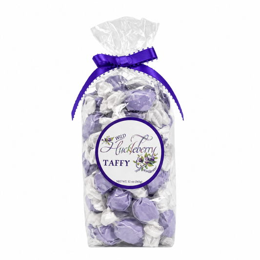 A clear cellophane bag filled with wrapped taffy candies in light purple and white, tied with a purple ribbon at the top. A round front label reads 'Wild Blueberry Taffy' with floral graphics.