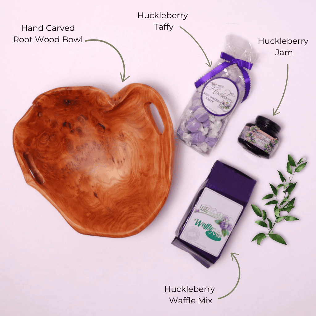 Flat Lay of Huckleberry Gift Basket Contents – A top-down view showcasing wild huckleberry jam, saltwater taffy, waffle mix, and a unique hand-carved wooden bowl, a perfect gourmet food gift.