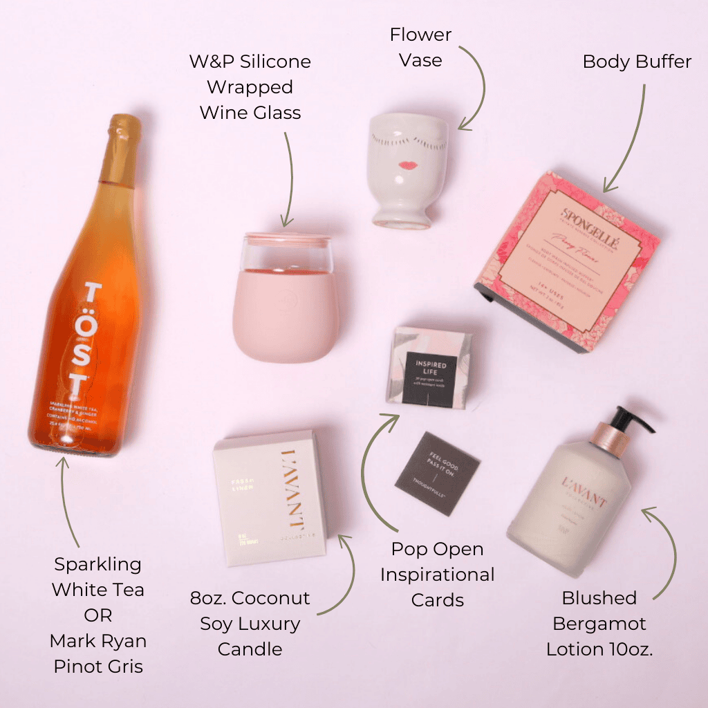 A beautifully curated luxury spa gift basket displayed in a flat lay, featuring TÖST Sparkling White Tea, L'AVANT Collective hand lotion, a fresh linen candle, a body buffer, a decorative vase, a glass wine cup, and uplifting ThoughtFulls pop-open cards.