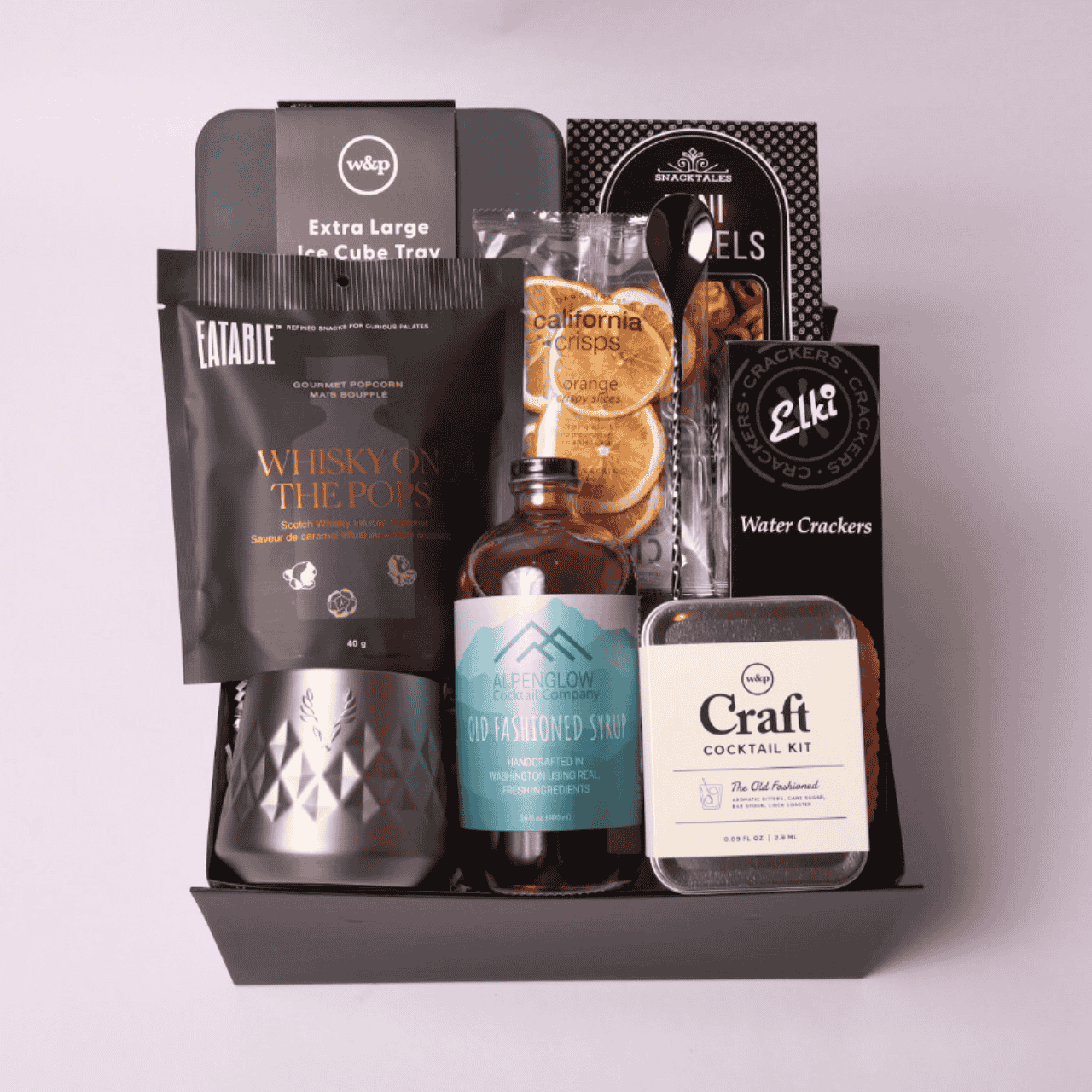 Sophisticated black gift box featuring cocktail-inspired treats: old fashioned cocktail syrup, whiskey tumbler, bar spoon, orange slices, pretzels, and crackers.