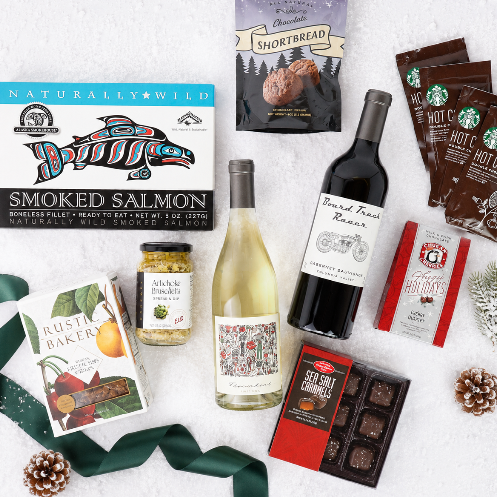 A curated gift basket flat lay on a white textured background, featuring a smoked salmon box, a bottle of white wine, a bottle of red wine, hot chocolate packs, a shortbread cookie bag, a box of Rustic Baker cookies, a jar of salami, chocolates, and a green ribbon.