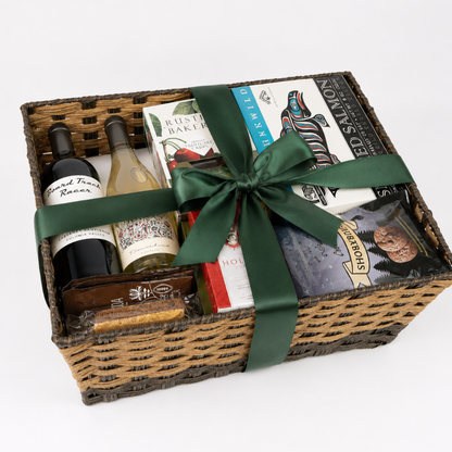 A wicker gift basket filled with assorted gourmet items, tied with a dark green ribbon. Visible contents include a bottle of red wine, a bottle of white wine, a box of rustic bakeries, a book, and packaged snacks.