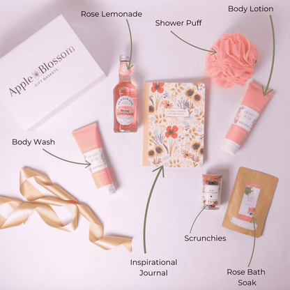 Flat Lay of Rose Garden Gift Box Contents – A beautifully arranged display featuring rose-scented body wash and lotion, Fentimans Rose Lemonade, a floral journal, mini scrunchies, a pink loofah, and a soothing tea sachet.