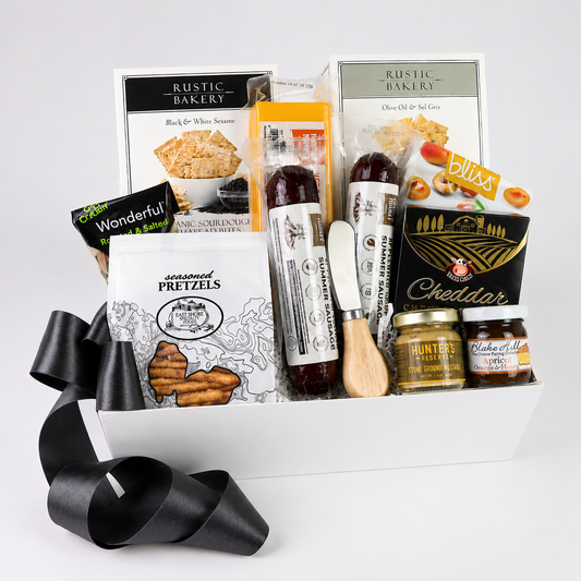 A curated gourmet gift basket from Rustic Bakery, filled with assorted cheeses, crackers, spreads, and a black ribbon bow, photographed on a clean white background.