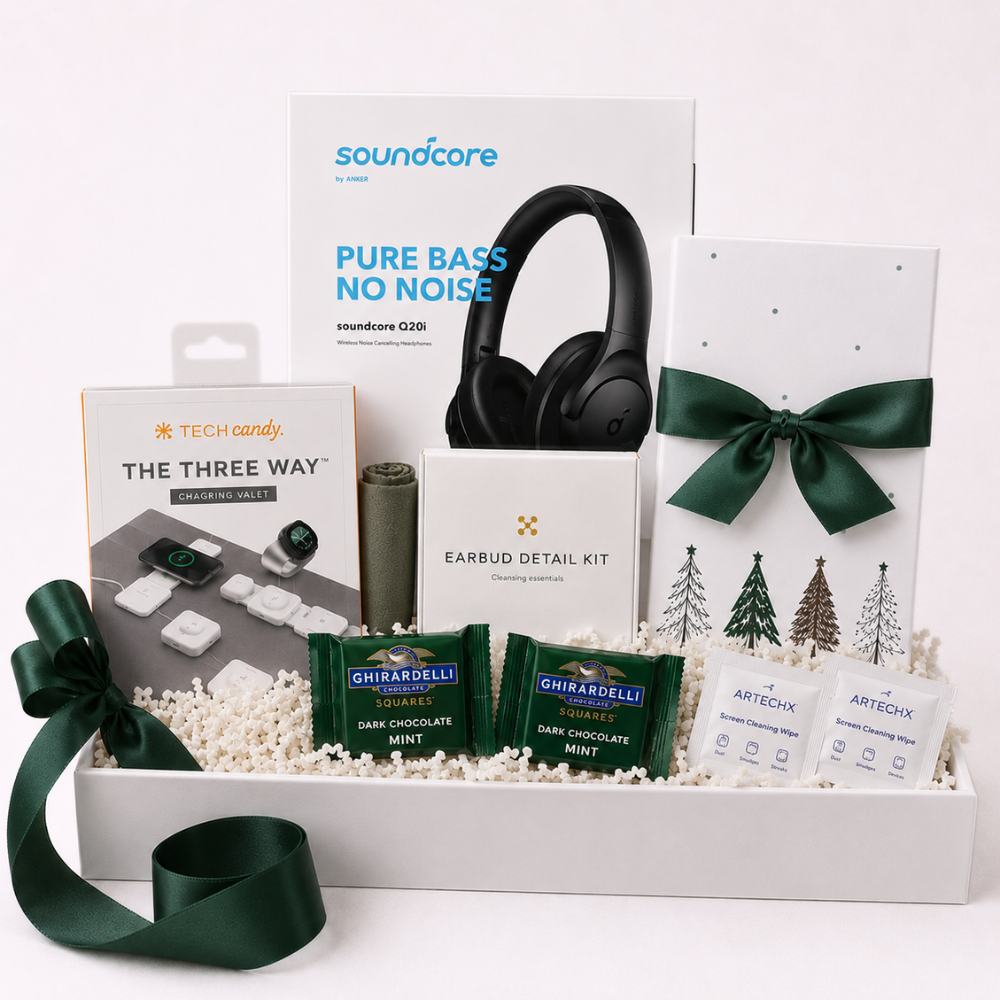 A curated gift box set on a white background, featuring a Soundcare headphones box, a Tech Candy 'The Three Way' game set, a green ribbon-wrapped box with Christmas tree graphics, earbud detail kit, Ghirardelli dark chocolate mints, and ArtEck packets arranged in white filler with a green ribbon bow.