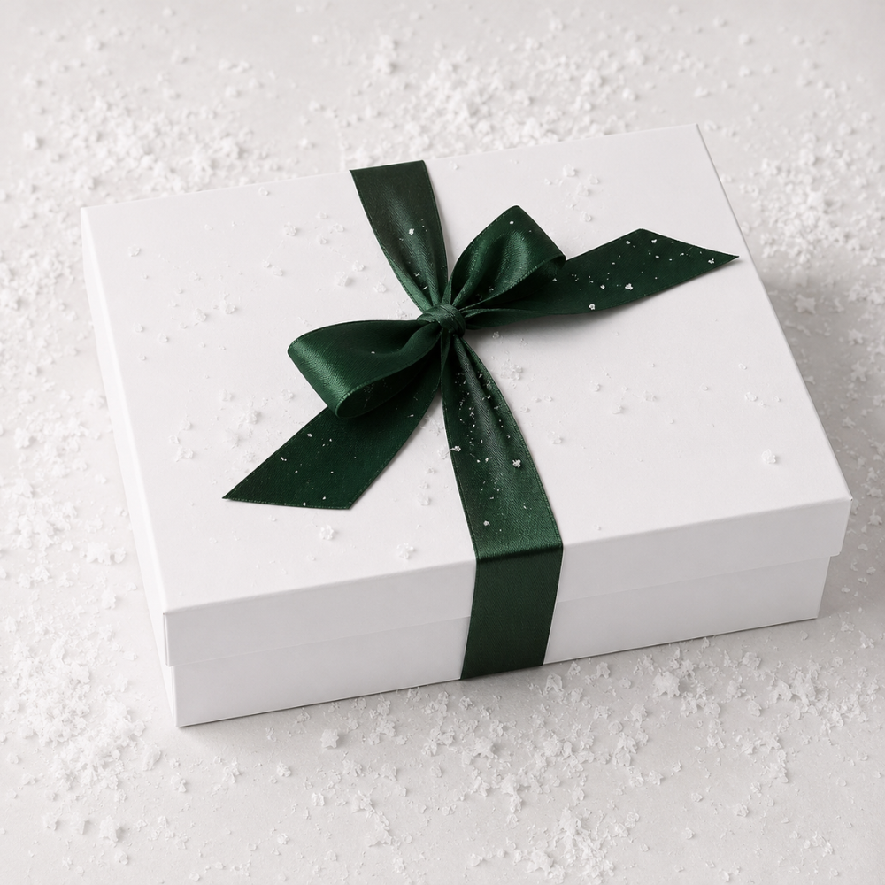 A white rectangular gift box tied with a dark green satin ribbon and bow, photographed on a light surface with scattered white specks.