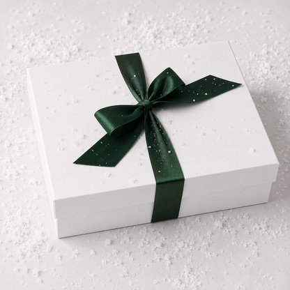A white rectangular gift box tied with a dark green satin ribbon and bow, photographed on a light surface with scattered white specks.