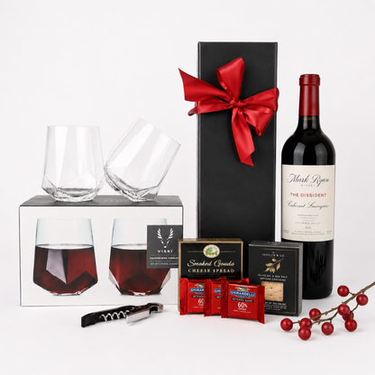 A curated gift set on a white studio background featuring a bottle of Mark ryan Red Label Winery wine, two faceted clear wine glasses in and out of their box, a tall black gift box tied with a red bow, smoked applewood cigar spruce boxes, red tea bags, a corkscrew, and red berries.