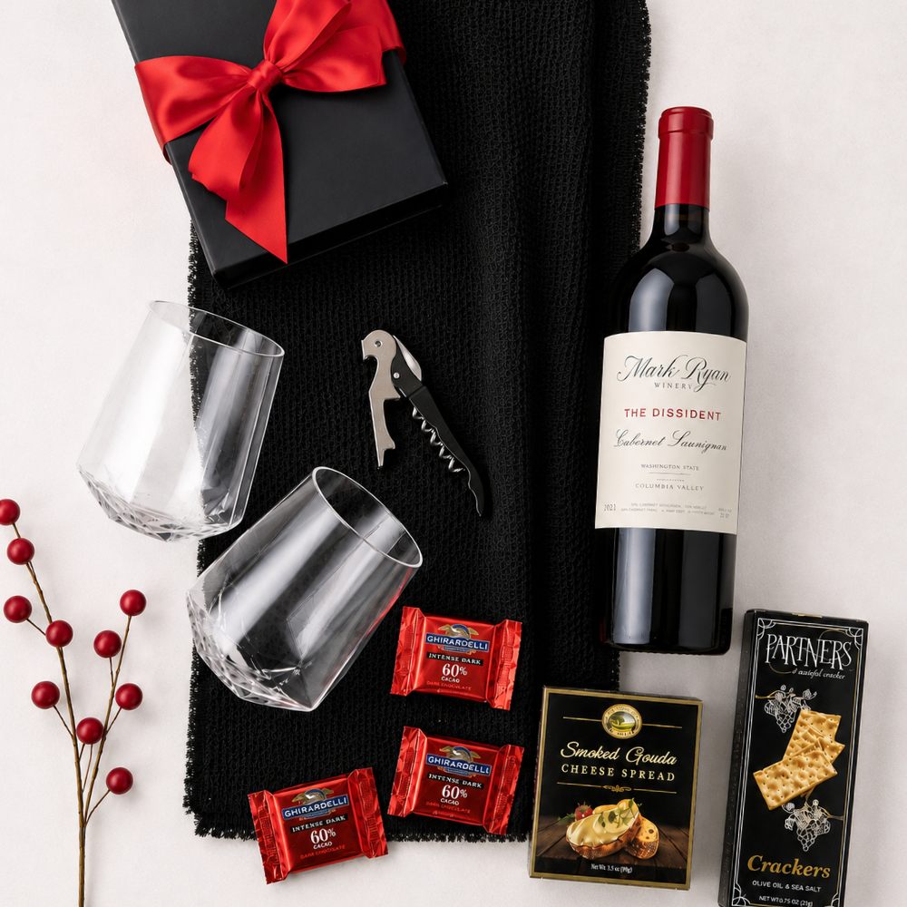 A gift basket arrangement on a light neutral background featuring a bottle of wine, two clear glasses, a corkscrew, red wrapped chocolates, crackers, cheese spread, a black gift box with a red bow, and a black textured cloth.