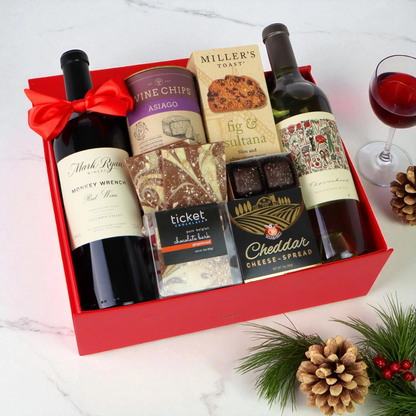 Gift box with wine bottles, cheese, and snacks on a white surface with a glass of red wine.