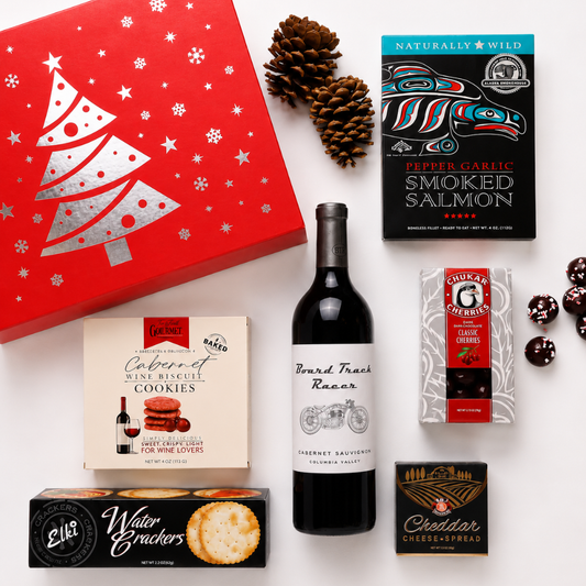 A curated gift assortment arranged on a white background, including a red Christmas-themed box, a bottle of red wine, a box of cookies, a pack of crackers, a package of wild salmon, and a box of cheese crackers, with pinecones as styling props.